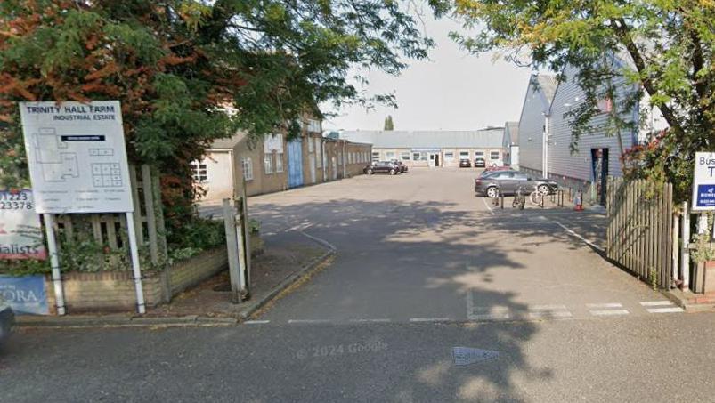 The entrance to Trinity Hall Farm Industrial Estate. On the left is a white sign, with its title at the top and underneath a map showing which unit is where. In the middle are the open gates and beyond it are single storey industrial units, metal clad on the right and brick built on the left. At the far-end is a single storey row of units. Cars are parked in front of some of the buildings. Trees are in leaf on either side of the gate. 