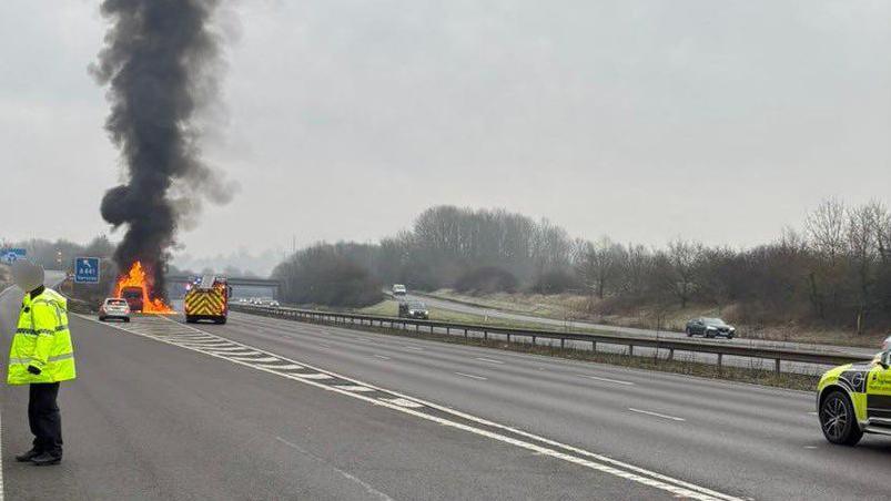 An image of a motorway junction, a van engulfed in flames pulled over in lane one, with a fire engine parked behind it. A person in a high-vis yellow jacket can be seen on the left and the front of a police car on the right. A few cars are driving on the other wide of the carriageway.