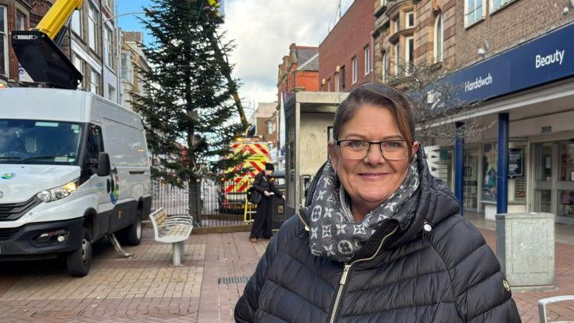 A woman smiling as she stands on Rhyl High Street in front of the tree. She is wearing a black padded coat and glasses.