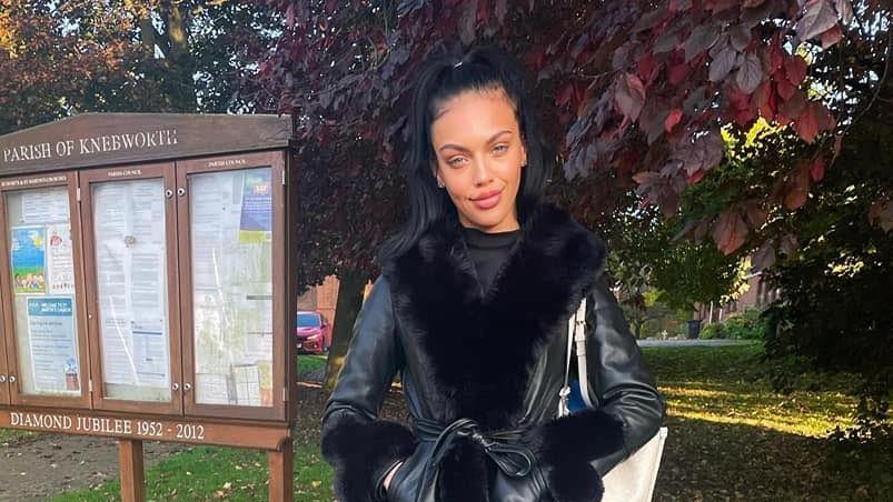 Bradford is standing outside next to a wooden noticeboard which reads 'Parish of Knebworth'. She is wearing a black coat with fluffy black accents on the ends of the sleeves and neck. She has long black hair in a ponytail and is smiling. There are trees behind her and leaves on the ground.