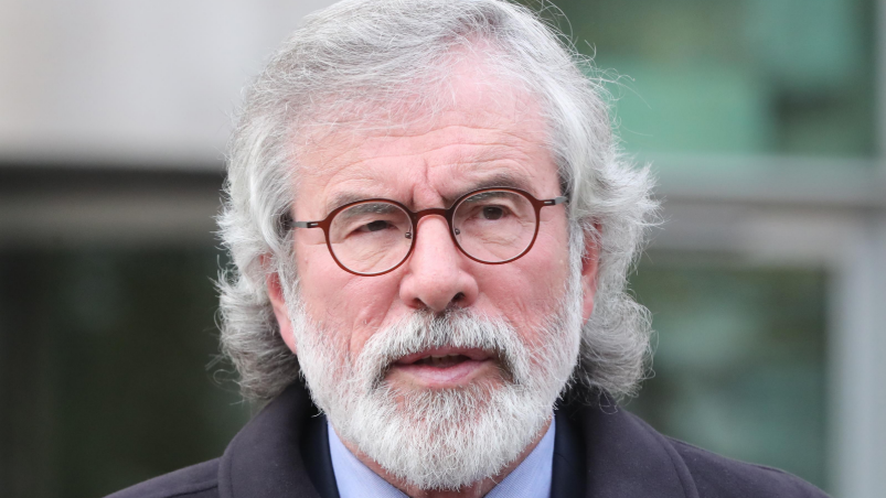 Gerry Adams is looking to the right of the camera. He has circular dark red glasses and a grey beard and hair down to his shoulder. 