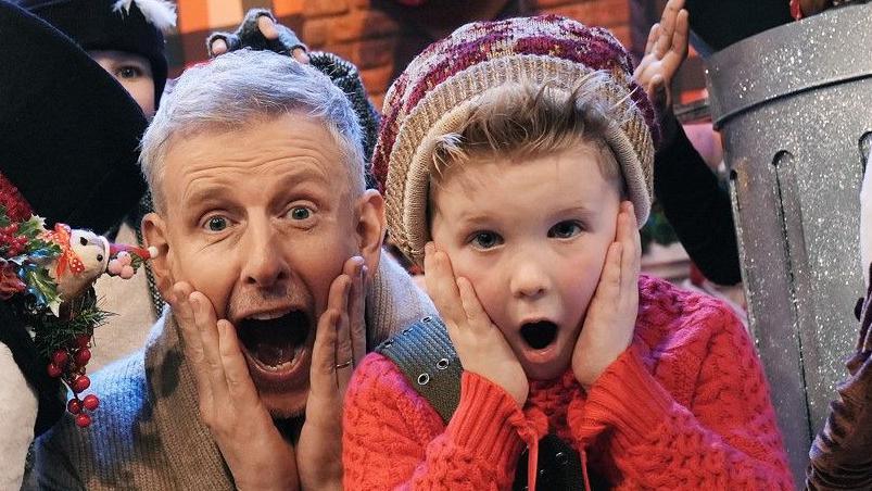 Patrick Kielty dressed in a Christmas cardigan with children. 
One is dressed as a snowman, one is dressed as a tree and one in a trash can.