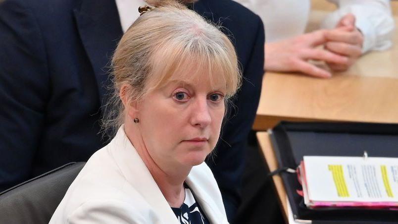 Shona Robison sitting at a desk looks at the camera side on, with a folder of paers on a desk in front of her