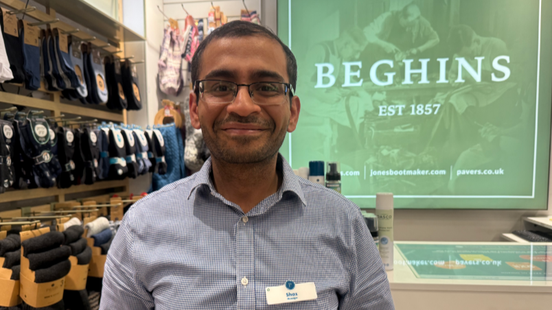A man wearing a striped shirt smiles into the camera. A Beghins sign is displayed behind him. Socks are display on a rail to the left.