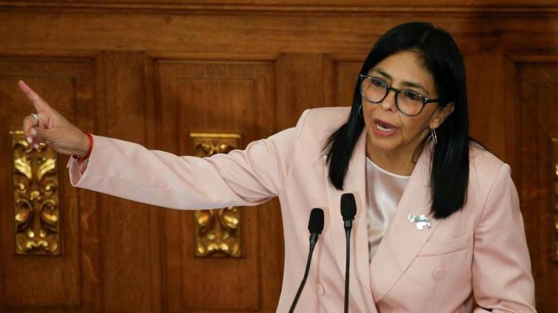 Venezuela's Vice-President Delcy Rodriguez presents the government's 2026 budget proposal to the National Assembly on 4 December 2025. She is wearing a salmon-coloured jacket and glasses. She is pointing with one hand and holding papers in the other. 