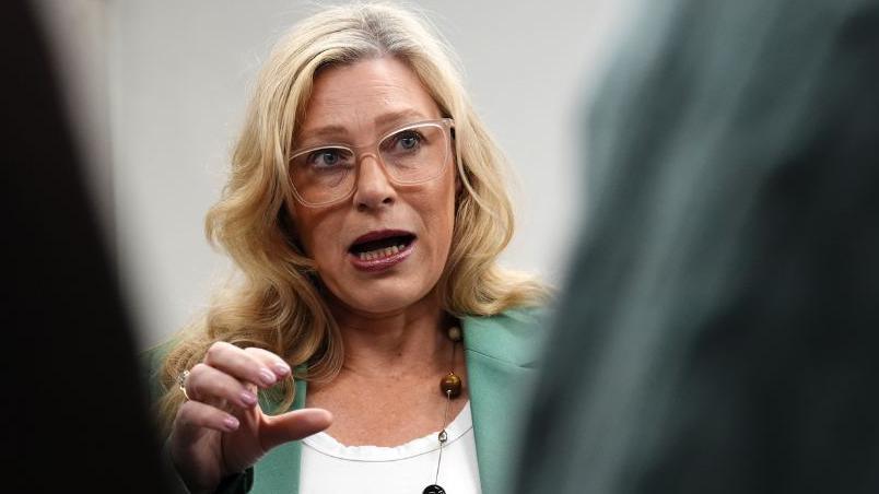 Gillian Martin, with blonde hair wearing a green suit and white top underneath, talks to the media in front of a grey background 