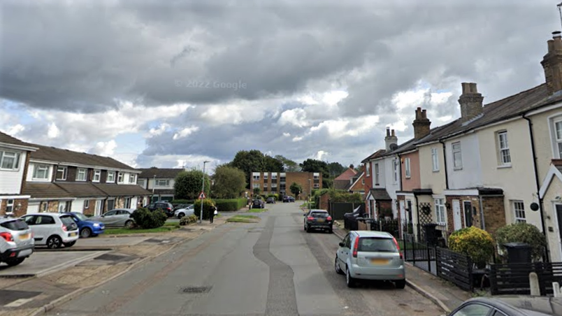 A road surrounded by multicoloured pastel terraced houses. There are black and grey cars parked on the right hand side of the road. 