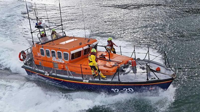 An orange and navy RNLI lifeboat with crew members standing on board as the boat cuts through the sea, causing white spray waves