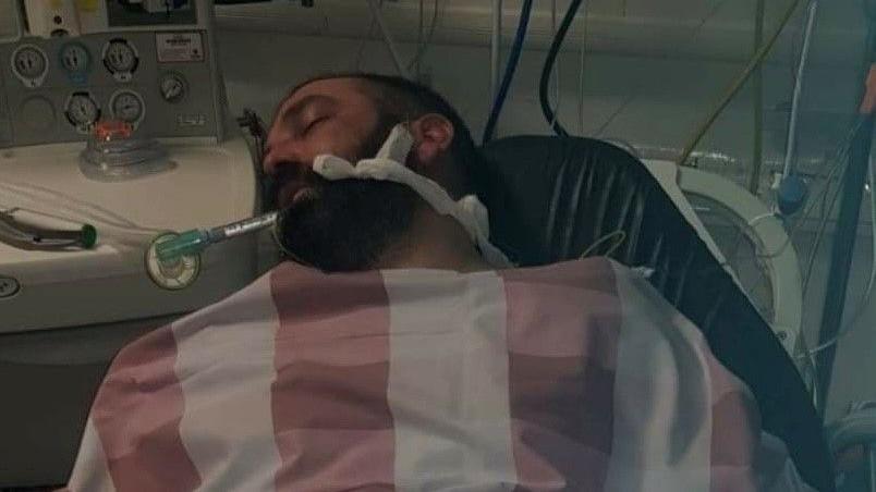 Scott Blackwell in a coma in a hospital bed, with a blanket over his chest and connected to various medical tubes