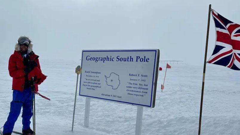 Capt Chandi in the South Pole. She is wearing a big read coat and is on skis. She is stood next to a sign which reads 'geographical South Pole'.