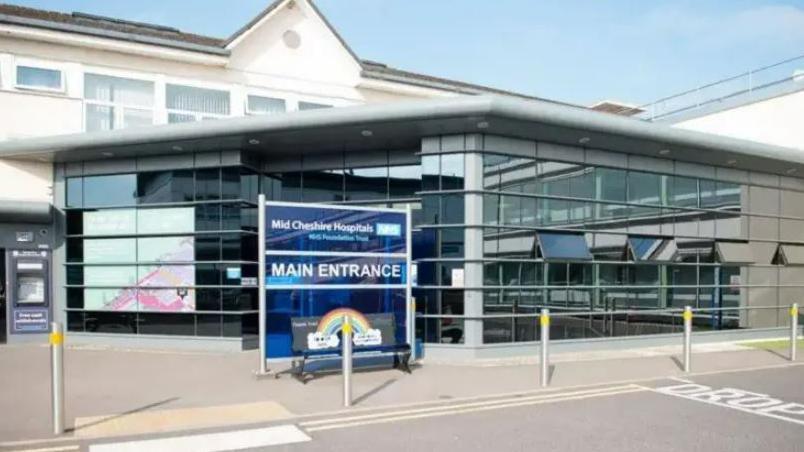 The entrance to Leighton Hospital, which has a large glass frontage, connected to a white building that can be seen behind it. A blue perspex sign is out the front, with Main Entrance written on it.