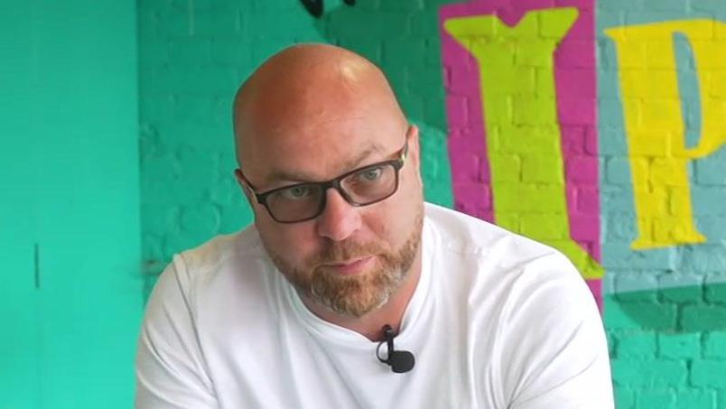 James Langan looks away from the camera. He is bald and has a light coloured beard and wears a white T-shirt with glasses. He stands in front of a green painted wall.