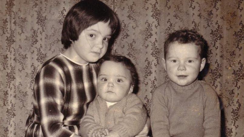A black and white photo of the three siblings sitting in front of a curtain, aged roughly between one years old and six years old.