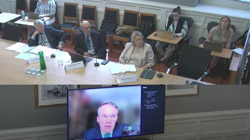 Prof Simon Mackenzie on a screen with a panel of three people sat at a table behind