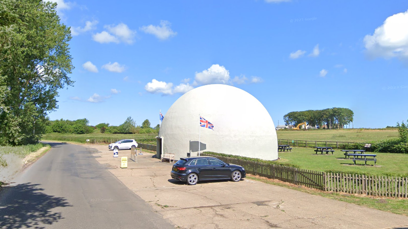 A white dome next to a road, with a Union Jack flag flying, cars parked and picnic tables on a grass area behind it