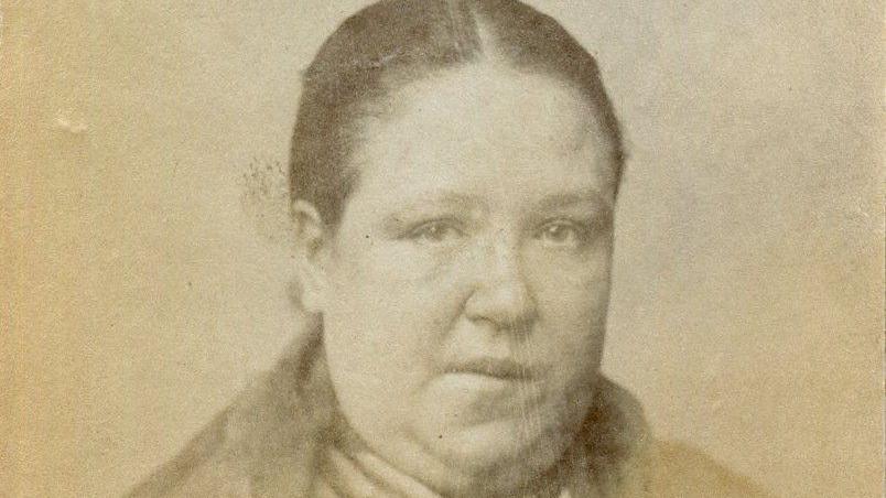A old sepia photograph of a woman sat with dark hair pulled back. He has a serious look on his face. 