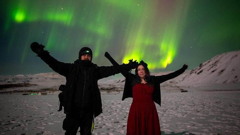 Simon Scott and Nicole Carr are standing in a snowy landscape and holding their arms in the air with the green Northern Lights above them.