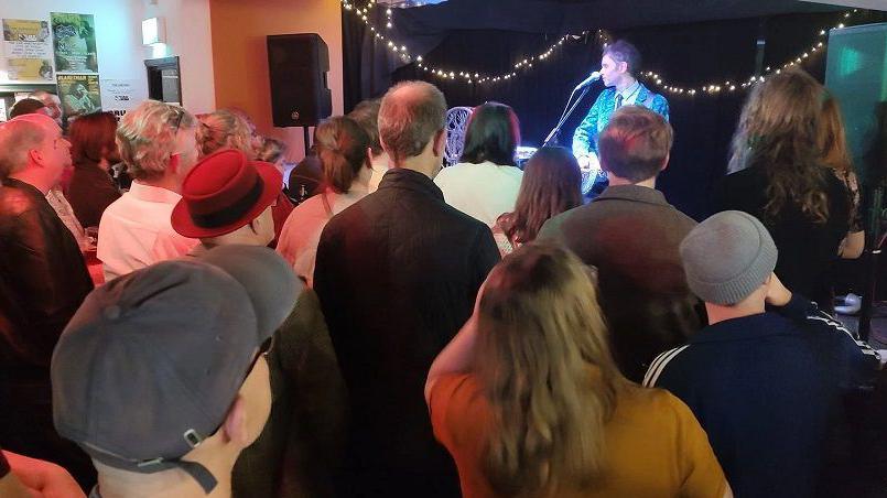 A crowd of people at an indoor gig watching a singer with a guitar