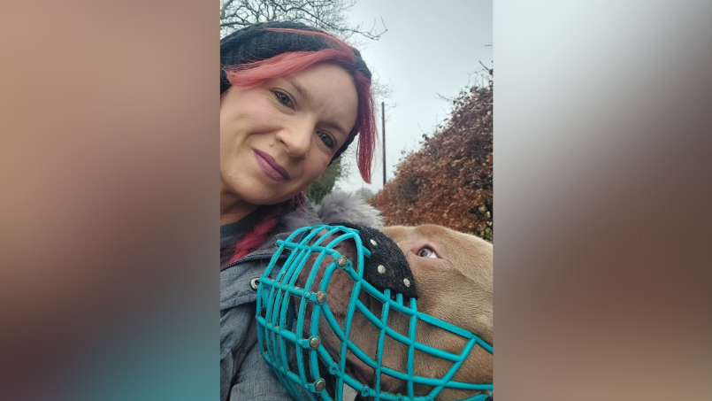 A selfie of Carla and Reggie while on a walk. Reggie is looking up at Carla with a turquoise muzzle on.  Carla has pink hair and is wearing a black hat and grey coat. She is smiling.