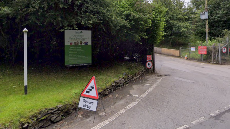 The entrance to the Crowndale Recycling Centre with a sign in the road saying queues are likely. There is a sign bearing the name of the centre and further writing which cannot be read in the image. There are also 5mph speed signs either side of the entrance.