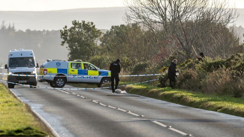 Police on road with police vehicles and police tape and officers looking in bushes