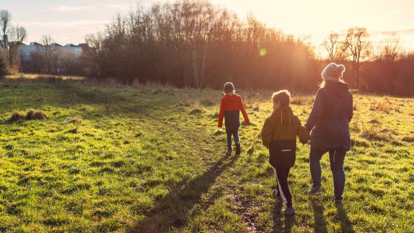 An adult woman and two children cast shadows in low winter light as they cross a field along the footpath.  The trees have lost their leaves and the sun is low in the sky.