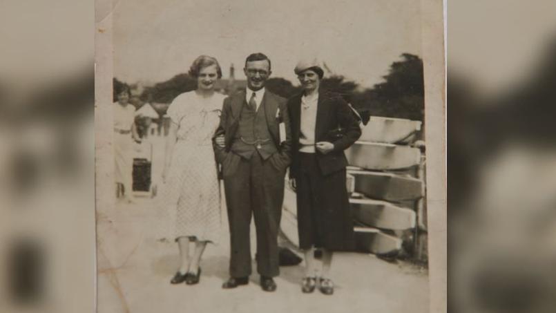 A black and white image of a young woman in a white dress standing next to a man in a three piece suit with a pocket watch, and a woman in a dark skirt and jacket with a white sweater and hat.