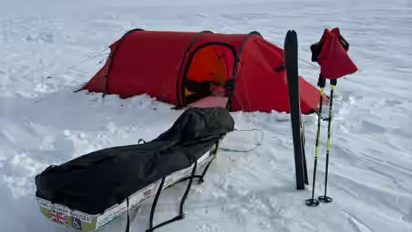 In the front there is a equipment on a sledge which can be dragged, ski's and  ski sticks. Behind there is a red tent. They is snow all around. 