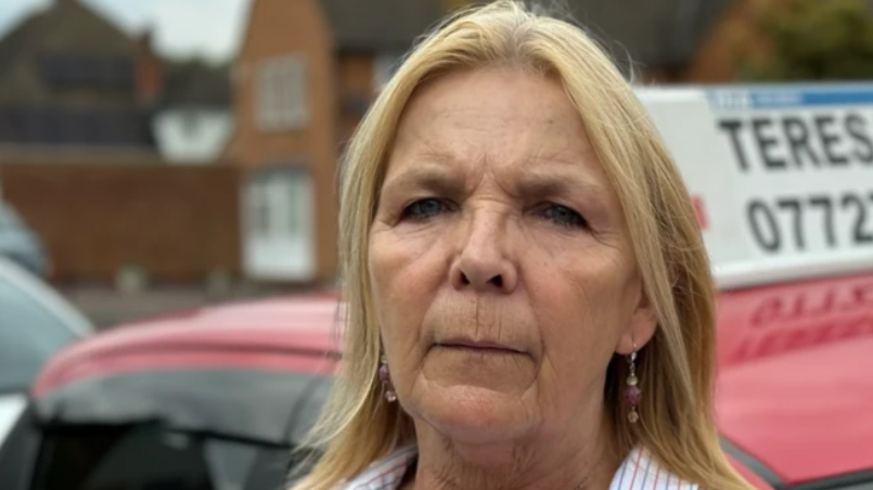 Teresa Allen is standing outside her red learner driver vehicle on a street in Kent. She has shoulder length blonde hair and is wearing a striped shirt, with a silver necklace