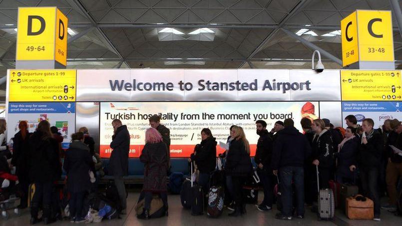 People queuing inside the terminal in London Stansted Airport. They are standing in front of a white board welcoming them to the airport, with yellow information boards offering directions.