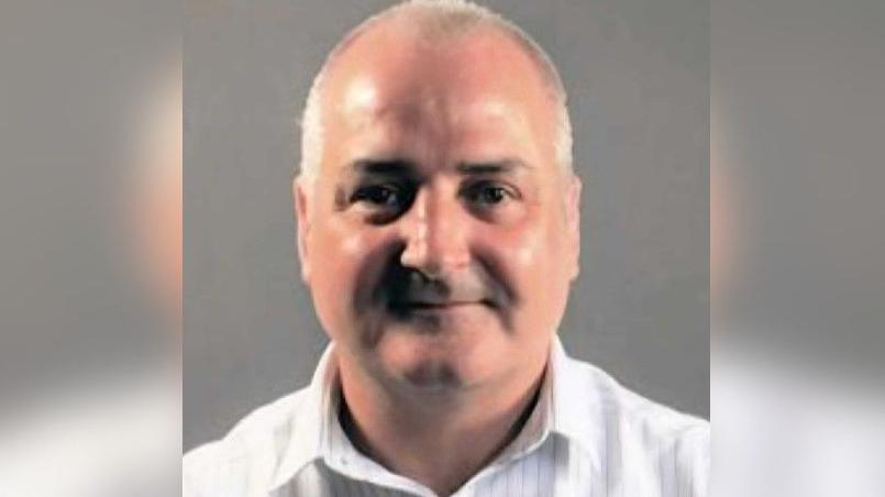 A head and shoulders shot of Sexton who is wearing an open neck white shirt. He is smiling and has very short grey hair.