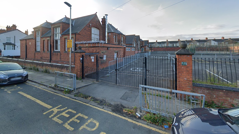 Brick school building with 5 windows facing a road and more windows along its side. There is a car park behind a brick wall with black gates and railings. There are two parked cars, weeds along a pavement and road with "Keep" written on it in yellow. There are houses in the distance. There is a lamp post and a yellow sign. There are also two grey railings next to the road.