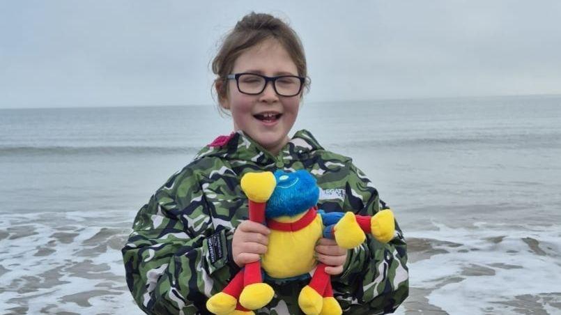 Lotus has brown hair and glasses and is wearing a camouflage coat and holding a bright yellow cuddly toy with a blue head and red legs. She is stood in front of the sea on a cloudy day