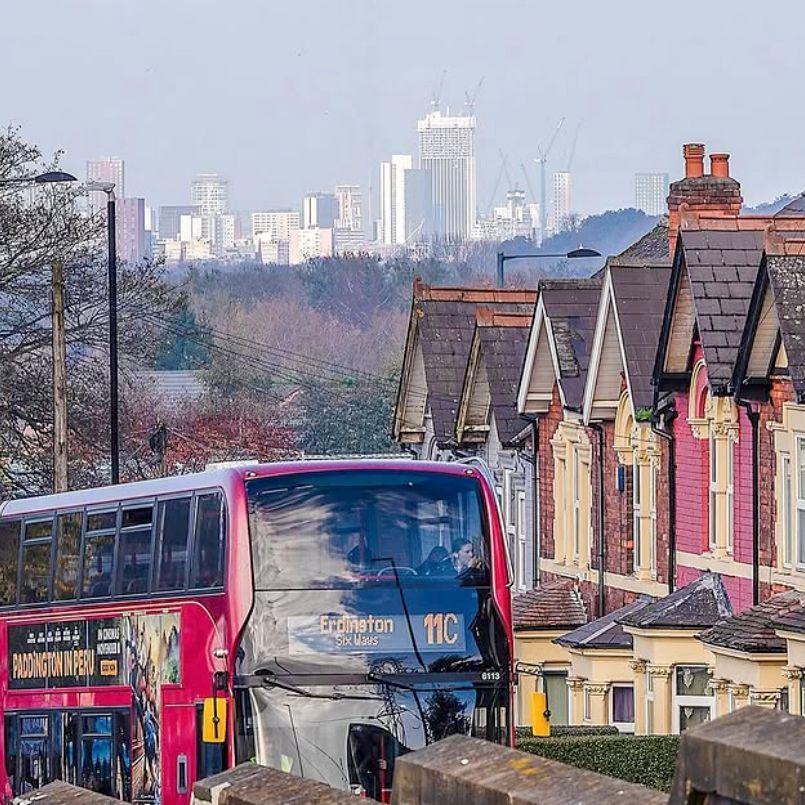 A red bus, saying 11C, travels past a row of terraced houses with a skyline of Birmingham city centre in the background. 