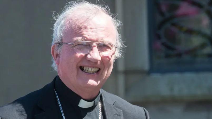 Donal McKeown is smiling at the camera. He is wearing a black jacket white and clerical collar with a large silver chain around his neck. He has white hair and gold glasses