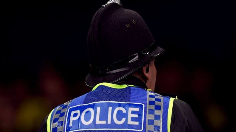 A close-up of the back of a police officer, who is wearing a blue and yellow trimmed vest with the word Police across it. They have a helmet on, and an earpiece in