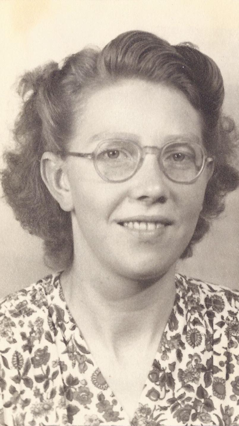 A sepia-toned photo of Mary Meijer. She is wearing spectacles and a floral top. The front and side sections of her short hair is pinned back and she is smiling at the camera.