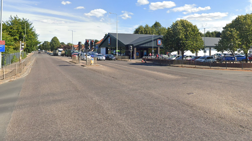 We can see a crossroads, from a view in the middle of the road. In the background we can see an Aldi supermarket.