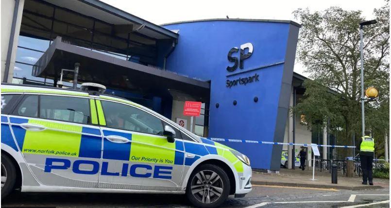 A police officer stands in front of police tape at the Sportspark, which is a large blue and white building. There is a police car at the front of the picture, also in front of the building.