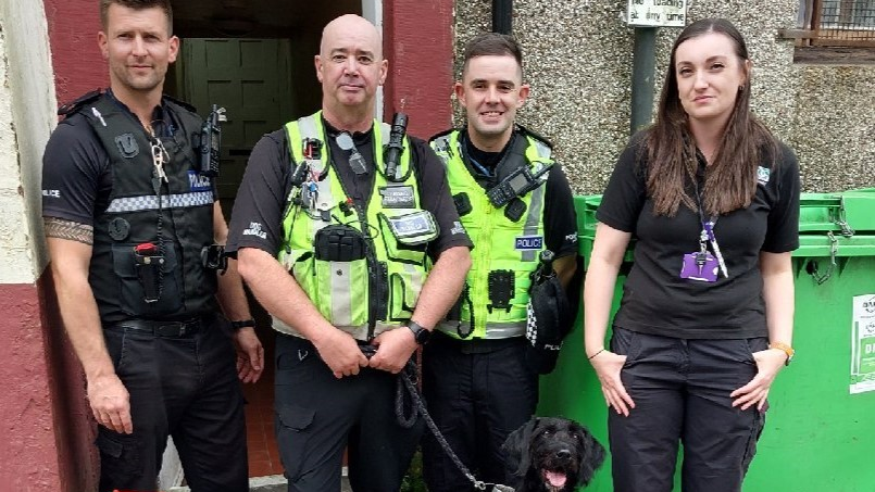 Police and trading standards officers wearing uniform stand in front of a doorway and a green wheelie bin. From left to right, PC David Steele, dog handler Bobby Cranie with Boo on lead, PC Stephen Dickson and Fiona Adrain.