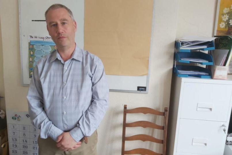 Man with a blue shirt standing with his hands folded in front of him. He is an office with a wooden chair and white filing cabinet.