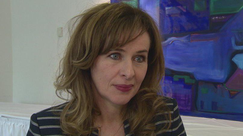 Prof Deirdre Heenan - a woman with brown hair wearing a black blazer jacket with white stripes stands in front of a abstract blue and green painting which is in a white room