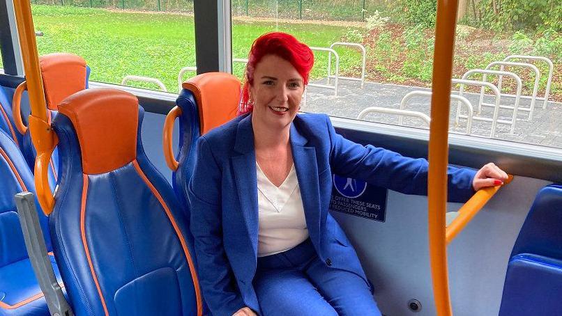 Louise Haigh smiles and looks at the camera as she sits on a blue bus seat. She is wearing a blue suit and has red hair. 