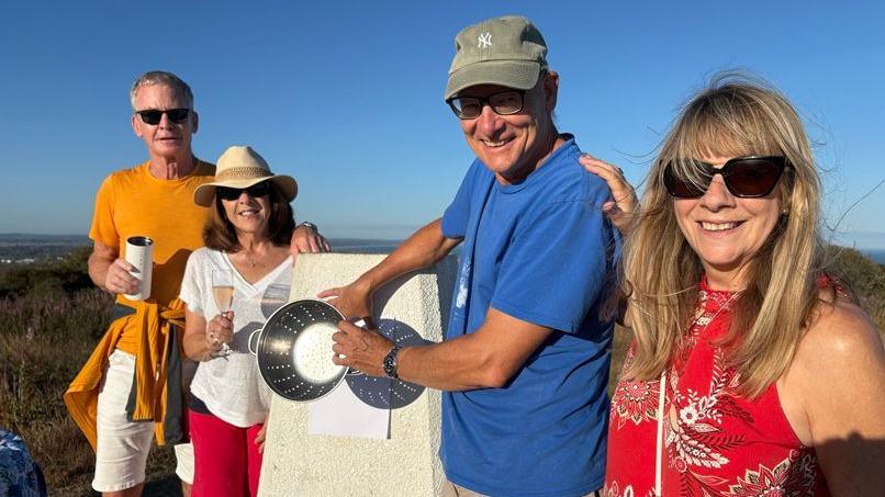 From left to right, Stephen Whiles, Glenda Whiles, David Hearn and Andrea Illingworth stand on the Eastbourne Downs during the eclipse. David Hearn holds a metal colander in front of a sheet of white paper to demonstrate a homemade viewing method, while Glenda Whiles holds a glass of sparkling wine. The group are gathered on open downland beneath a clear evening sky.