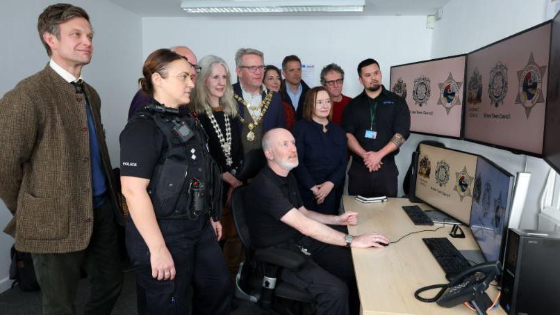 A group of people stand at a CCTV desk which is being controlled by a man. They are all looking towards a bank of screens above the desk. Members of the group include councillors and police.