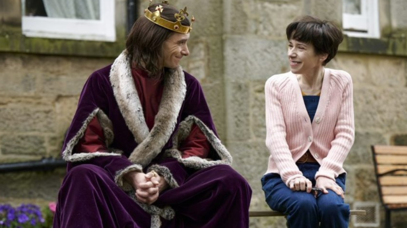 Sally Hawkins, wearing a pink cardigan and jeans, sitting next to an actor in a royal cloak and crown.
