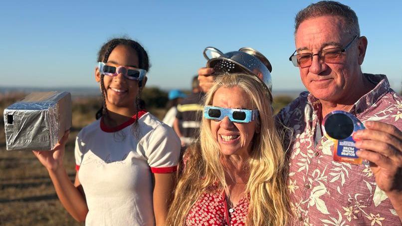 From left to right, Chloe Simpson, Anne Simpson and Mike Simpson stand on the Eastbourne Downs wearing eclipse glasses. Chloe Simpson holds a homemade eclipse viewer covered in silver foil, while Mike Simpson holds a pair of eclipse-viewing filters. Other spectators can be seen in the background watching the event.