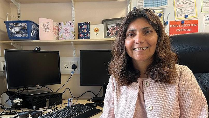 GP Pallavi Devulapalli is standing in South West Norfolk for the Green Party. She is sitting in her consulting room wearing a light pink jacket. 