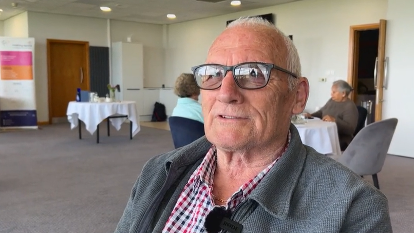 An elderly man wearing black-rimmed glasses and a checked shirt, sitting down