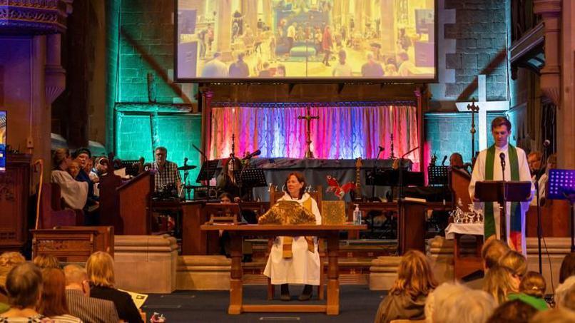 Church with video screen in centre, above altar (on which stands a wooden cross). To the right, the vicar with short dark hair is standing in front of a music stand wearing a white cassock and a green stole. The bishop with long dark hair, wearing a white cassock and a yellow and red stole, is setting at a table in the centre. There are various musicians seated behind them and audience members in front
